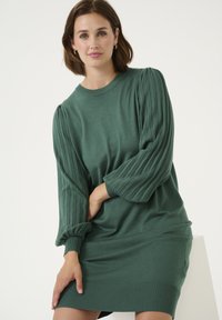 Robe en tricot vert avec de longues manches plissées et des poignets côtelés. Texture douce, encolure ronde et coupe décontractée.