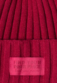 Rode gebreide beanie met ribbelstructuur en een stoffen label met de opdruk "VIND JE INNERE VREDE" in geëmbossde letters.