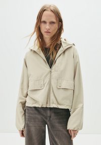 Beige, leichte Zip-Jacke mit Kapuze, die zwei Fronttaschen und einen elastischen Bund aufweist. Kombiniert mit grauen Jeans.
