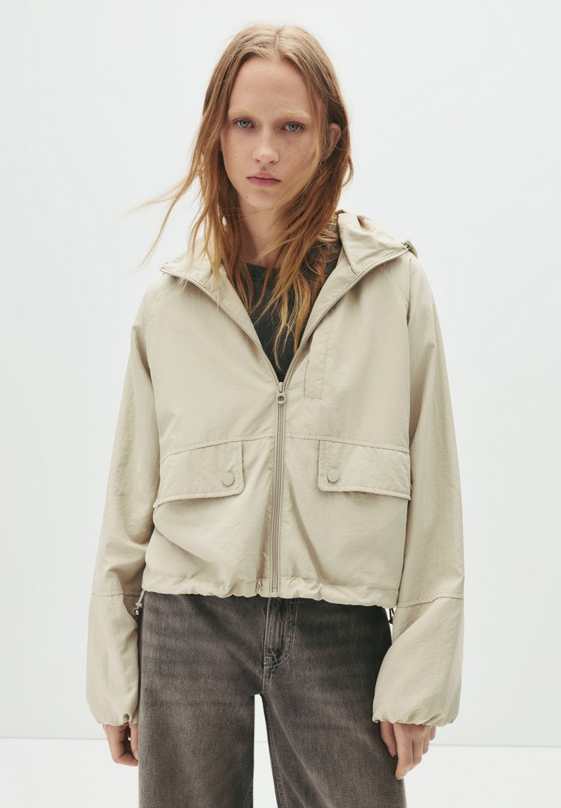Beige, leichte Zip-Jacke mit Kapuze, die zwei Fronttaschen und einen elastischen Bund aufweist. Kombiniert mit grauen Jeans.