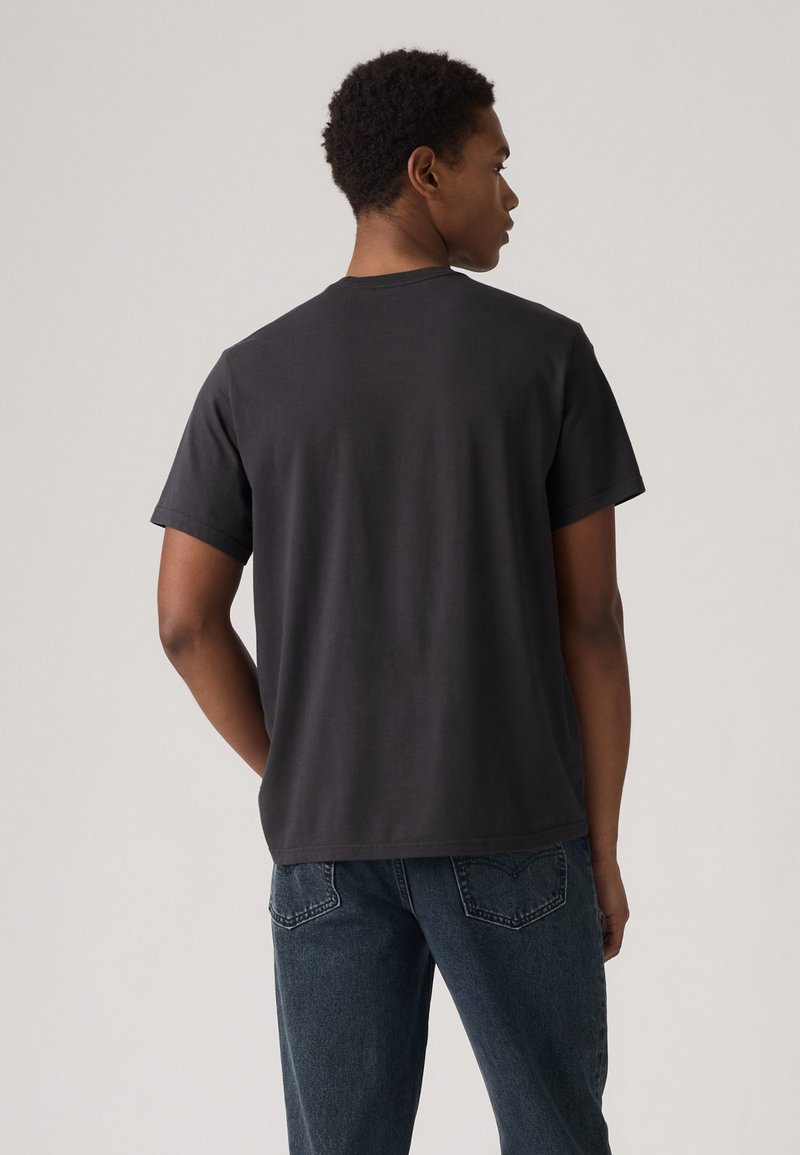 T-shirt gris foncé à manches courtes et col rond, en coton. Le dos est uni, sans logos ni motifs visibles. Un jean en denim est porté.