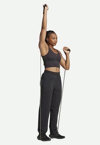 Juodas sportinis liemenėlis ir laisvi juodi džinsai, laikantys rezistencijos juostas, pakelta ranka, sportiniai bateliai, neutralus fonas.