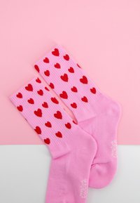 Różowe skarpetki z czerwonymi wzorami serc położone płasko na dwukolorowym różowo-białym tle.