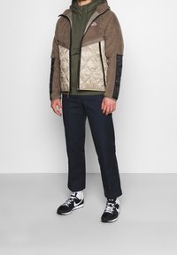 Brun och beige hybridjacka med plyschig textur, vadderade sektioner, svarta tygdetaljer, bärs över en olivgrön hoodie och mörka byxor.