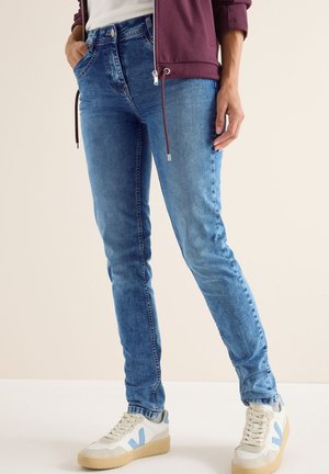 Los pantalones vaqueros ajustados de denim azul presentan un clásico diseño de cinco bolsillos con cierre de cremallera y botón. Combinados con zapatillas blancas acentuadas en azul.