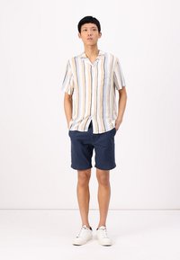 Camisa de manga corta con rayas verticales en beige y azul, combinada con pantalones cortos marinos y zapatillas blancas. Tejido suave, corte relajado y detalle de bolsillo.