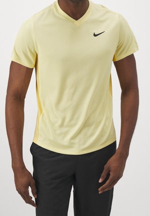 Mann trägt ein gelbes Nike-V-Ausschnitt-T-Shirt mit kurzen Ärmeln und schwarze Shorts, steht vor einem weißen Hintergrund.