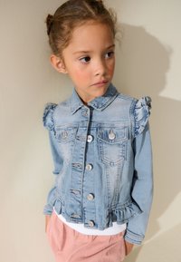 Junges Mädchen mit hellbraunen Haaren in zwei Dutts, das eine blaue Jeansjacke mit Rüschen und eine rosa Hose trägt und seitlich an eine beige Wand schaut.