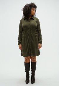 Olívazöld gombos ingruha puha anyagból, hosszú ujjakkal, gallérral és combközépig érő hosszúsággal. Térdig érő fekete csizmával párosítva.