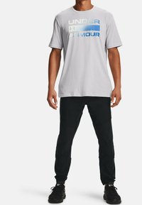 Camiseta de algodón gris con "UNDER ARMOUR" impreso en un degradado de azul y blanco. Combinada con pantalones negros y zapatillas deportivas negras.