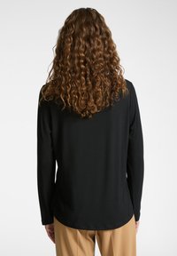 Schwarzes Langarmtop mit abgerundetem Saum, aus weichem Stoff gefertigt. Hat eine glatte Textur und ein einfaches Design, kombiniert mit beigen Hosen.