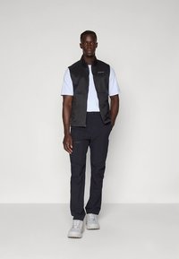 Black sleeveless vest over a white t-shirt, paired with dark pants and light-colored shoes. Smooth textures and minimal design details.