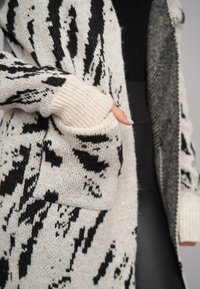 Sweter kardigan z czarno-białym abstrakcyjnym wzorem, o luźnym kroju, z bocznymi kieszeniami i ściągaczami przy mankietach. Miękka textura i ciepły materiał.