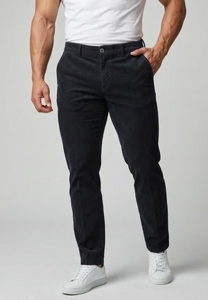 Mann trägt eng anliegende schwarze Hose, weiße Sneakers und ein weißes T-Shirt mit einer Hand in der Tasche vor schlichten Hintergrund.