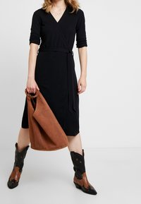 Černé šaty s dlouhými rukávy a páskem na zavazování, zkombinované s hnědou semišovou kabelkou a černými kovbojskými botami se hvězdnými detaily.