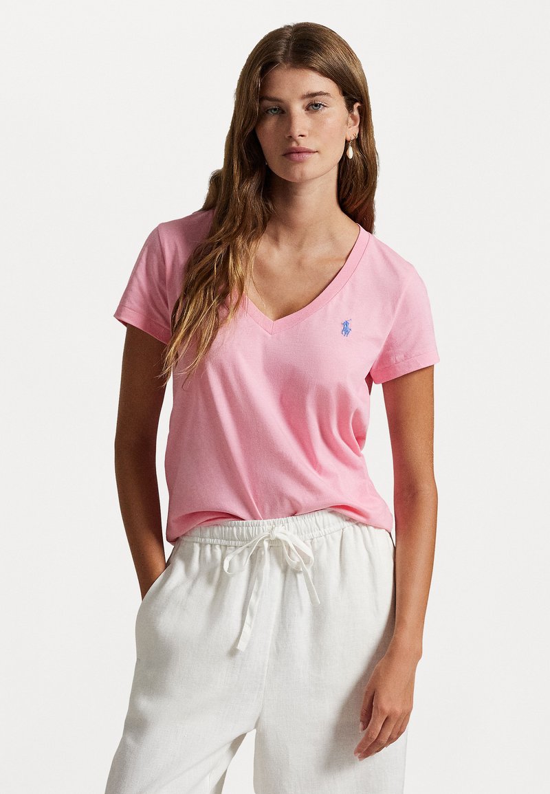 T-shirt en coton rose avec un col en V, des manches courtes et un petit logo bleu sur la poitrine gauche ; associé à un pantalon blanc léger et ample.