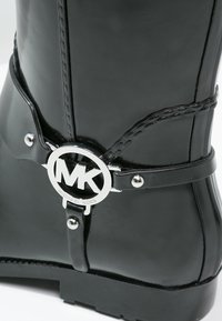 Czarne gumowe buty z srebnym logo "MK" przymocowanym ozdobnym paskiem. Gładka tekstura, okrągły nosek i masywna podeszwa.