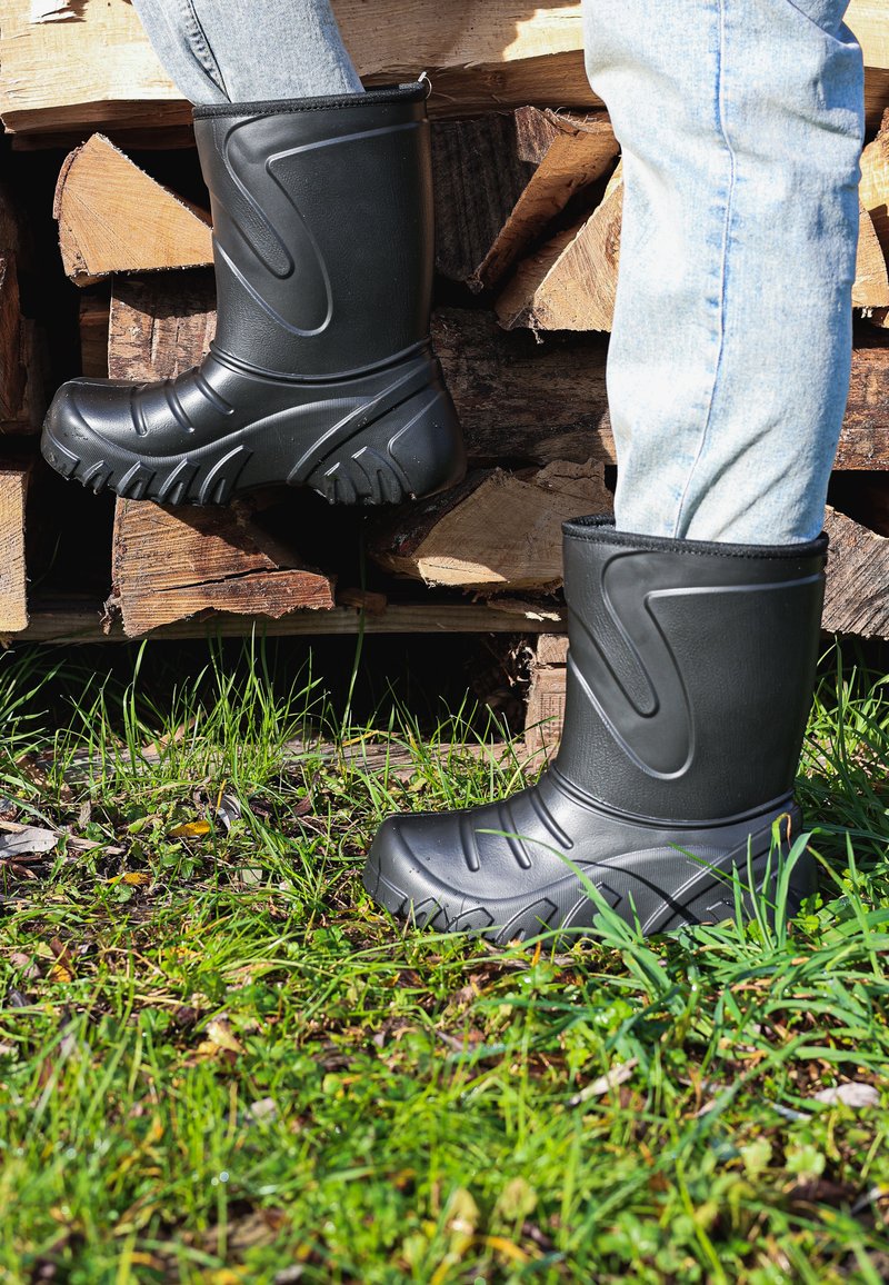 Zwarte rubberen laarzen met een gestructureerd oppervlak, een gebogen ontwerp en een grove zool, geplaatst tegen gras en houten stammen.
