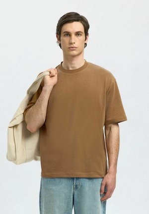 Bruine katoenen T-shirt met een ronde hals en korte mouwen, gecombineerd met lichtblauwe denim jeans. Model houdt een crèmekleurige jas over één arm.