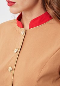 Mujer con chaqueta beige con botones dorados, ribete dorado y collar mandarina rojo, mostrando los labios y parte de la cara y el cabello.