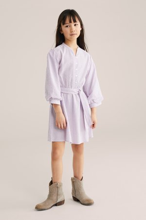 Meisje met lang donker haar, gekleed in een lichtpaarse gestreepte jurk met knopen en een riem, en beige enkellaarsjes, staand tegen een effen achtergrond.