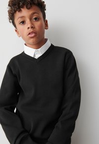 Črni pulover z V-izrezom in dolgimi rokavi, nošen čez belo srajco z ovratnikom. Gladek tekstura, minimalističen dizajn, brez vidnih vzorcev ali poudarkov.