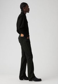 Zwarte overhemd met knopen, donker groene spijkerbroek met een leren patch aan de achterkant, en zwarte westernlaarzen. Zijaanzicht.