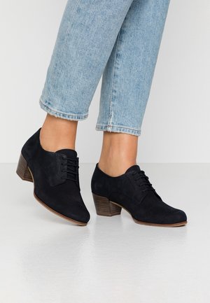 Woman wearing light blue jeans and black low-heeled lace-up shoes standing on a reflective white floor.