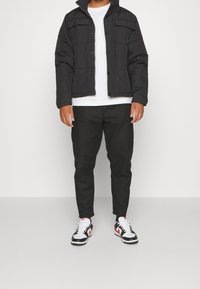 Man som står och bär en svart vadderad jacka, vit t-shirt, svarta byxor och vita sneakers med svarta och röda detaljer.
