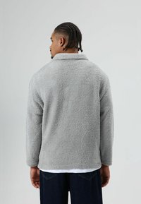 Grijze fleecejas met een zachte, gestructureerde afwerking, voorzien van een kraag en lange mouwen, gedragen over een wit shirt.