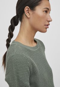 Pull en maille verte avec un motif texturé, col rond et accents côtelés au niveau du col. Le modèle a une queue de cheval tressée.
