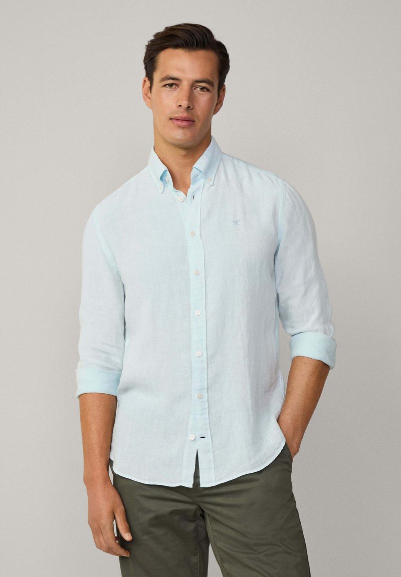 Camisa de lino azul claro con cuello abotonado, mangas largas remangadas y un pequeño logo bordado en el lado izquierdo del pecho.