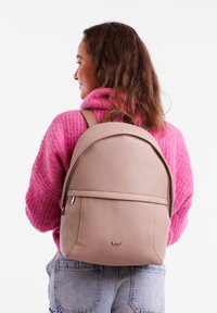 Beige leather backpack with a rounded shape, textured surface, and zippered front pocket, worn over a pink sweater, with adjustable straps.