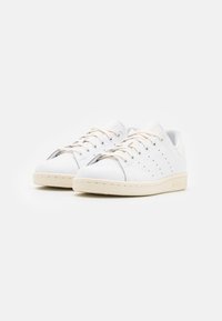 Zapatillas de piel blancas con lados perforados, cordones planos y suela beige. Punta redondeada y diseño minimalista.