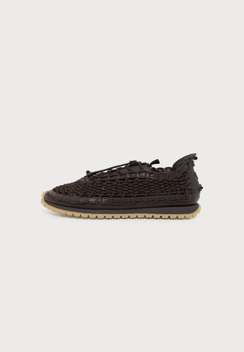 Sneaker tissé marron foncé à maille ouverte avec semelle en caoutchouc beige et languette noire sur fond blanc uni.
