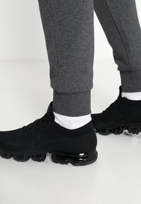 Svarta sportskor med övre del i mesh, som har en texturerad design och reflekterande detaljer, i kombination med grå joggers och vita strumpor.