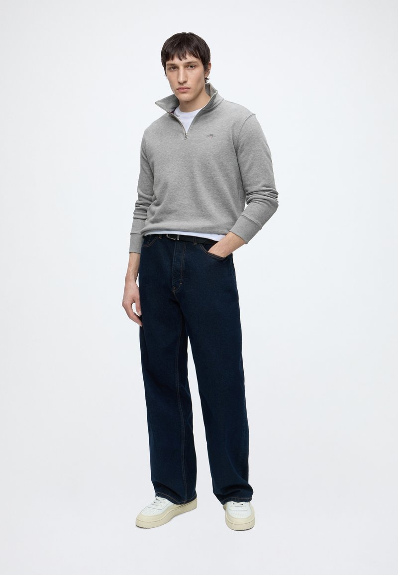 Junger Mann, der mit einer Hand in der Tasche steht, trägt einen grauen Viertel-Reißverschluss-Pullover, dunkle weit geschnittene Jeans und weiße Turnschuhe vor einem einfarbigen Hintergrund.