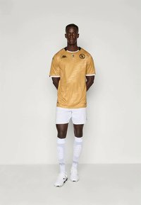 Gul fodboldtrøje med korte ærmer, sort logo og detaljer. Sammen med hvide shorts og knælange sokker med prikket mønster.