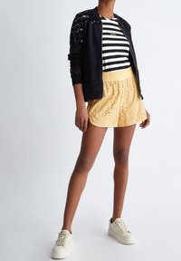 Černá bomber bunda s flitrovými rameny, pruhované tričko a žluté šortky s potiskem, doplněné bílými teniskami.