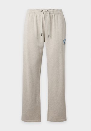 Homme aux cheveux courts noirs, debout, portant un t-shirt à manches courtes gris clair et un pantalon de survêtement assorti avec des baskets blanches sur un fond uni.