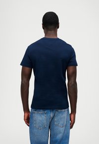 Ναυτικό μπλε βαμβακερό T-shirt με κοντά μανίκια και V-λαιμόκοψη, συνδυασμένο με ανοιχτό μπλε τζην παντελόνι, εμφανίζεται από πίσω.