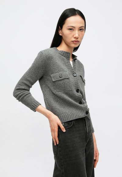 Γυναίκα που φοράει γκρι ζακέτα με κουμπιά και τσέπες με καπάκι, συνδυασμένη με σκούρο τζιν, στέκεται μπροστά σε ένα απλό φωτεινό φόντο.