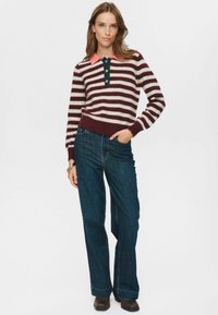 Frau steht mit der Hand in der Tasche, trägt einen bordeaux-rot-weißen gestreiften Pullover mit einem pinken Kragen, dunkelblaue Jeans und braune Stiefel.