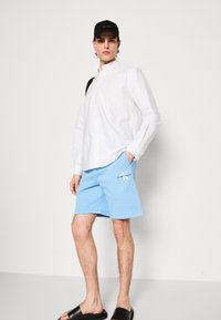 White button-up shirt, light blue shorts with logo, black cap, and black sandals. Casual outfit with a relaxed fit and minimal details.