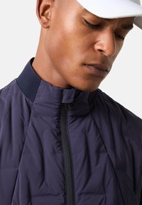 Navy lightweight jacket with a high collar, a zippered front, and textured accents. Smooth, puffy fabric with a modern fit.