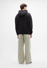 Felpa nera oversize con cappuccio trapuntato, abbinata a pantaloni in denim larghi di colore verde chiaro, dotati di tasche posteriori e cuciture a vista.