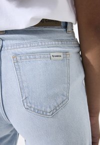 Jeans de mezclilla azul claro con un parche en el bolsillo trasero etiquetado como "GARCI" y detalles de costuras visibles. La tela tiene una textura suave.