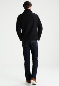 Svart ulljacka med fleecekrage, i kombination med mörkblå jeans och bruna sneakers, visad bakifrån.