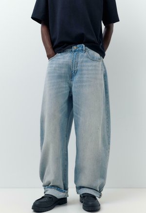 Person iført lyseblå vidde jeans, sorte sko og en mørk, kortærmet skjorte med hænderne i lommerne, stående mod en ensfarvet baggrund.