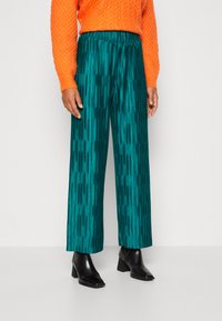 Pantalones de pierna ancha en color teal con un acabado texturizado, combinados con un suéter de punto trenzado naranja y botines negros con tacón cuadrado.